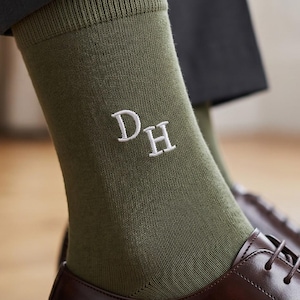 May include: Olive green crew-length socks featuring white embroidered initials DH on the side. The socks are worn with dark grey trousers and polished dark brown leather oxford shoes, showcasing a ribbed cuff and a smooth knit texture.
