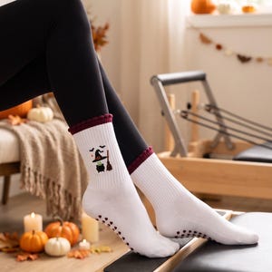 May include: White crew socks with a ribbed texture and a dark red cuff. The socks feature a small graphic of a black witch hat, a bubbling green cauldron, and a broomstick. The soles of the socks have black silicone grip dots for traction.