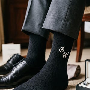 May include: A pair of black crew-length socks featuring a subtle diamond-knit texture and the white embroidered initials BW on the outer side. The socks are worn with grey dress trousers and black leather dress shoes.
