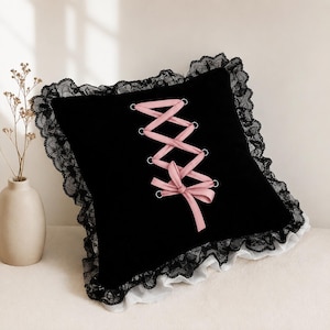 May include: A square black velvet throw pillow featuring a central pink ribbon corset-style lace-up design with silver metal eyelets. The edges are trimmed with a ruffled black lace border layered over a white fabric ruffle base.