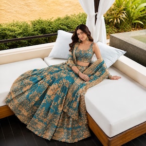 Könnte beinhalten: Ein blaugrünes Lehenga-Set bestehend aus einem ärmellosen bauchfreien Oberteil und einem voluminösen bodenlangen Rock, beide mit aufwendigen goldenen Blumenstickereien und Pailletten verziert. Ein passender transparenter Dupatta mit breitem Goldrand.