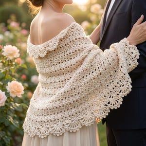 May include: An off-the-shoulder cream-coloured crochet shawl featuring an intricate openwork pattern and a scalloped edge. The garment drapes over the shoulders and arms, showcasing a detailed yarn texture with a repeating decorative border.