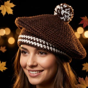 May include: A dark brown crocheted beret featuring a white and brown striped band around the base and a matching white and brown pom-pom on top. The hat is crafted with a textured yarn stitch pattern and has a rounded, structured shape.