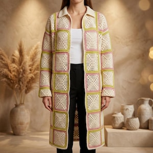 May include: A long-sleeved, knee-length crochet cardigan featuring a grid of square motifs in cream, light green, and soft pink. The garment has a collared neckline, an open front, and is worn over a plain white top and black trousers.