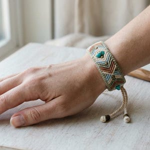 May include: A woven macrame bracelet featuring a geometric pattern in shades of beige, light blue, and rust orange. The bracelet is accented with small metallic beads and two heart-shaped turquoise stones, finished with adjustable braided cord ties.