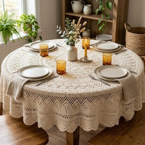 Puede incluir: Una mesa de comedor redonda cubierta con un mantel de ganchillo de color crema con patrones intrincados. La mesa está puesta con cuatro platos blancos, cuatro vasos de color ámbar y servilletas de lino. Un jarrón con flores secas está en el centro.