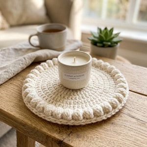 May include: A round, cream-colored crocheted coaster with a textured bobble-stitch border. Centered on the coaster is a white ceramic candle jar with a label that reads SUNDAY MORNING AMBER & OAK, featuring a single visible cotton wick.