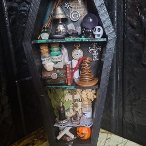 Op de afbeelding: Een zwart doodskistvormig houten rek met drie niveaus met miniatuurvoorwerpen, waaronder glazen toverdrankflesjes, een zilveren lamp, een kristallen bol, een schedel, een zakhorloge, een bruine puntmuts, een metalen kruis, een sleutel en een pompoen.
