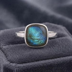 May include: A silver ring featuring a square-shaped labradorite gemstone with a smooth, polished surface. The stone displays iridescent blue and green flashes across its face. The band is a simple, polished silver metal with a rounded profile.
