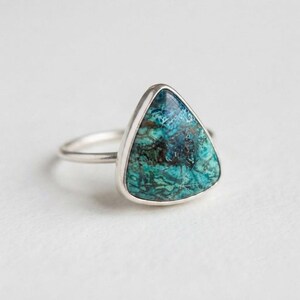 Pode incluir: Um anel de prata com uma pedra de formato triangular apresentando um padrão marmorizado de veios em azul-petróleo profundo, turquesa e castanho escuro. A banda de metal é fina e polida, segurando a pedra num engaste liso e simples.