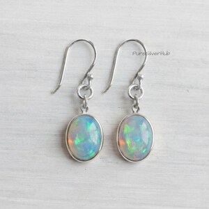 Puede incluir: Un par de pendientes colgantes de plata con ópalos ovalados que muestran destellos iridiscentes en verde, azul y naranja. Las piedras están engastadas en biseles de plata lisa y cuelgan de ganchos sencillos. El texto PureSilverHub es visible.