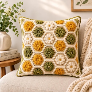 May include: A square throw pillow featuring a crochet pattern of interconnected hexagonal motifs. Each hexagon contains a raised floral design in shades of mustard yellow, olive green, and cream white, bordered by a cream-colored yarn frame.