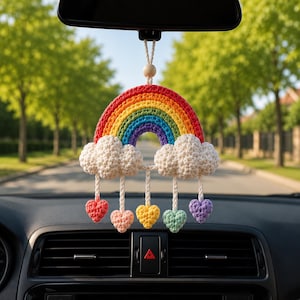 May include: A crocheted rainbow car accessory featuring a vibrant red, orange, yellow, green, blue, and purple arch. Two white crocheted clouds are attached to the base of the rainbow, with five small crocheted hearts in various colors hanging below.