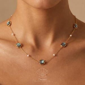 May include: A gold chain necklace featuring five square-shaped iridescent abalone shell charms and small white pearls spaced evenly along the links. The necklace rests against the neck, showcasing a delicate design with polished metal settings.