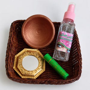 Op de afbeelding: Een donkerbruine gevlochten mand met een terracotta kom, een goudkleurige achthoekige spiegel met versierde randen, een groene cilindrische tube met gouden bloemmotieven en een doorzichtige spuitfles met een roze dop en een oog op het etiket.