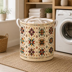 May include: A cylindrical laundry hamper with a cream-colored quilted exterior featuring a repeating geometric star pattern in shades of red, blue, green, and purple. The hamper includes two white side handles and is filled with a soft white fabric.