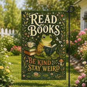 May include: A rectangular garden flag with a dark green background featuring a frog reading a book. The text reads READ BOOKS BE KIND STAY WEIRD. The design includes a stack of books, a teacup, a dragonfly, flowers, and a mushroom in a forest setting.