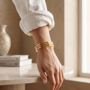 Könnte beinhalten: Ein goldfarbenes Armband in Manschettenform, bestehend aus drei parallelen, dünnen und glatten Reifen, die das Handgelenk umschließen. Das Armband hat eine polierte, reflektierende Oberfläche und ein schlichtes, offenes Design.