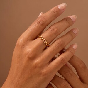 May include: A gold ring featuring a continuous chain link design. The band is composed of small, interconnected oval-shaped metal loops that create a textured, repeating pattern around the entire circumference of the jewelry piece.