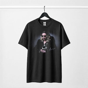 May include: A black short-sleeve t-shirt featuring a graphic print of a person with long hair and sunglasses wearing a dark jacket over a patterned shirt. The print includes a signature-style script text below the figure on the chest area.