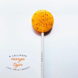 May include: A round yellow mango-flavored lollipop with red chili flakes embedded in the surface, attached to a white stick printed with the text LECCARE LOLLIPOPS. Below, text reads 6 LOLLIPOPS mango tajin LICK SMILE REPEAT with small red hearts.