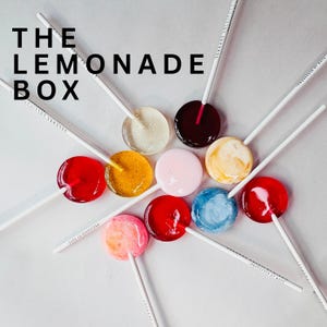 May include: An overhead shot showcases a collection of colorful lollipops on white sticks, arranged in a circular pattern. The lollipops are various colors, including red, yellow, pink, and blue. The text "THE LEMONADE BOX" is displayed in black at the top left.