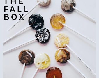 May include: A collection of round lollipops on white sticks arranged on a white surface. The candies feature various colors including black, brown, gold, and cream with marbled or speckled textures. The text THE FALL BOX is visible.