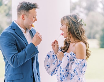 May include: A blue suit jacket worn over a white collared shirt and an off-the-shoulder long-sleeved blouse featuring a white base with a blue floral pattern, both individuals holding and eating small lollipops on thin sticks.