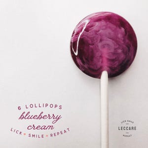 May include: A purple and white swirl lollipop on a white stick. The lollipop is labeled "6 lollipops blueberry cream lick smile repeat" and "lick smile leccaree repeat".