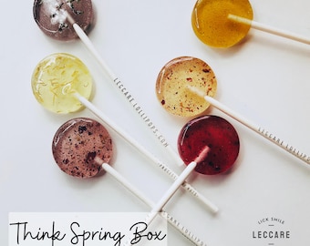 May include: Assortment of six colorful lollipops with different floral and herbal inclusions. The lollipops are arranged on a white surface with the text "Think Spring Box" and "Leccare Lollipops.com" printed on the sticks.