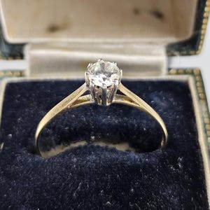 Könnte beinhalten: Ein Goldring mit einem einzelnen runden, klaren Edelstein in einer silberfarbenen Fassung mit sechs Krappen. Der Ring befindet sich in einer dunkelgrünen, mit Samt ausgekleideten Schatulle, auf deren Innendeckel NORTHERN GOLDSMITHS COMPANY NEWCASTLE-ON-TYNE steht.