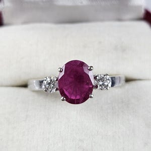 May include: A silver-toned ring featuring a large, oval-cut, deep pinkish-red gemstone at the centre, flanked by two smaller, round-cut, clear white stones. The ring is set in a polished metal band and presented inside a velvet-lined jewellery box.