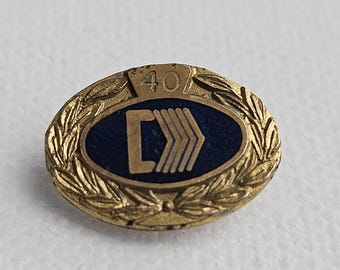 Pode incluir: Um broche oval de metal dourado com um centro azul escuro apresentando uma letra C dourada e quatro listras em chevron. A borda consiste em um design de coroa de louros texturizada, com o número 40 em relevo no topo da moldura.