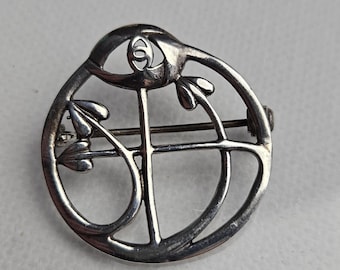 Puede incluir: Un broche circular de tono plateado con un diseño calado, una barra vertical central y líneas curvas que forman un motivo floral estilizado. La pieza incluye un pequeño emblema circular con un logotipo en la parte superior y un alfiler horizontal.
