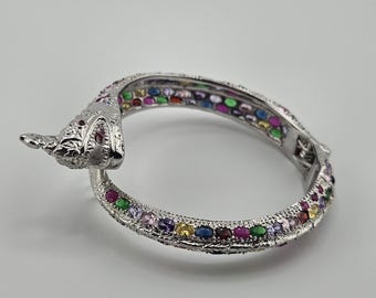 Pode incluir: Uma pulseira rígida articulada em forma de cobra de tom prateado com uma superfície metálica texturizada e uma série de pedras ovais multicoloridas em tons de vermelho, verde, azul, amarelo e roxo incrustadas ao longo da banda.