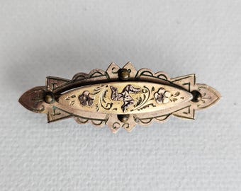 Pode incluir: Um broche de metal dourado com uma placa central oval alongada, apresentando padrões florais gravados e um símbolo de triskelion no centro. O broche possui uma moldura decorativa recortada com pequenos detalhes esféricos nos pontos cardeais.