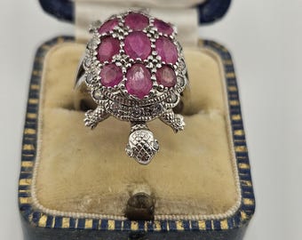 Pode incluir: Um anel prateado em forma de tartaruga, com um casco adornado com várias pedras preciosas ovais de cor vermelho-rosado e pequenas pedras claras ao longo da cabeça, patas e borda, apresentado dentro de uma caixa de joias azul forrada.