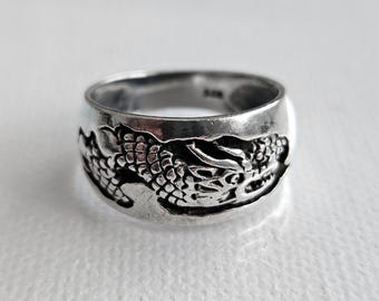 Pode incluir: Um anel de metal prateado com um design detalhado de dragão em relevo que envolve a banda. A superfície do anel tem um acabamento polido com áreas rebaixadas escurecidas para destacar as escamas e os traços do dragão.