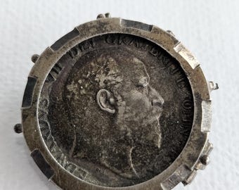 Pode incluir: Um broche de moeda circular em tom prateado com um retrato de perfil de um monarca barbudo voltado para a direita. A moeda está inserida numa moldura metálica com pequenos pinos salientes ao longo da borda externa e texto latino gravado.