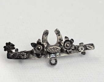 Pode incluir: Um broche de metal prateado com um motivo central de ferradura ladeado por duas flores de cinco pétalas. A barra horizontal é decorada com pequenos detalhes florais adicionais, folhas e um pequeno laço circular pendurado no centro.