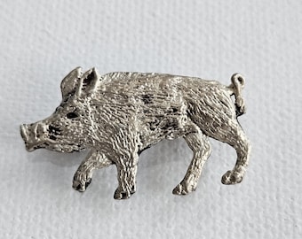 Pode incluir: Um broche de metal prateado em forma de javali. A peça apresenta uma superfície texturizada que imita a aparência de pelo áspero, com um focinho pontiagudo, orelhas eretas e uma pequena cauda enrolada, capturado de perfil em posição de andar.