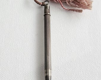 Pode incluir: Uma lapiseira metálica com um corpo texturizado de design entrecruzado. O topo da lapiseira está preso a um laço de cordão trançado que se conecta a uma borla decorativa rosa empoeirada feita de fios finos e soltos.