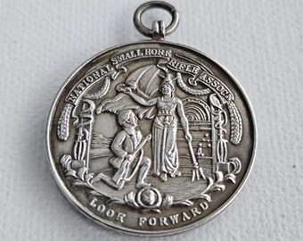 Pode incluir: Uma medalha circular de metal prateado com um design em relevo de uma figura ajoelhada e uma figura em pé segurando um tridente. O texto NATIONAL SMALL BORE RIFLE ASSOCN está arqueado no topo, com LOOK FORWARD inscrito na parte inferior.