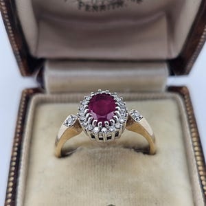 May include: A gold ring featuring an oval-cut deep red ruby center stone surrounded by a halo of small round diamonds. The band is crafted from yellow gold and includes additional small diamonds set into the shoulders of the ring setting.