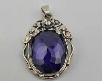 Pode incluir: Um pingente oval com uma grande pedra preciosa facetada de cor roxa profunda, engastada numa moldura de prata. A estrutura de metal é decorada com motivos florais e de folhas que rodeiam a pedra central, com uma argola de prata.