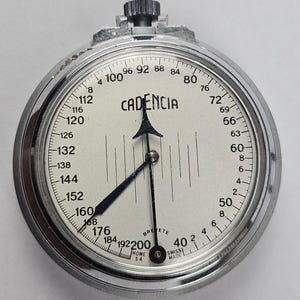 May include: A silver-toned Cadencia stopwatch with a cream-colored dial. The face features black numerical markings ranging from 4 to 200, vertical line indicators, and the text 'CADENCIA', 'BREVETE', 'HOME SA', and 'SWISS MADE' in black ink.