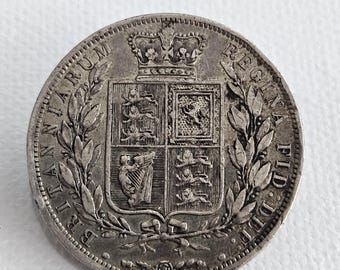 Pode incluir: Uma moeda circular de cor prateada com um escudo central dividido em quatro quadrantes com leões e uma harpa. Uma coroa situa-se sobre o escudo, rodeado por uma coroa de louros. O texto BRITANNIARUM REGINA FID DEF está gravado na borda.