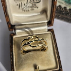 Puede incluir: Un anillo de oro con un diseño de banda ondulada de varias capas con tres pequeñas piedras redondas y transparentes incrustadas a lo largo de las curvas superiores. El anillo se presenta dentro de un joyero marrón con bisagras.