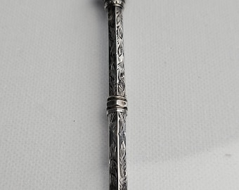 Pode incluir: Um estojo para agulhas em metal prateado com um corpo hexagonal alongado e gravações florais intrincadas. O topo é rematado com um acabamento decorativo texturizado e o corpo é dividido por uma banda central, exibindo padrões detalhados de folhas.