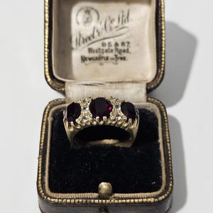 Op de afbeelding: Een gouden ring met drie ovaal geslepen donkerrode stenen en kleine heldere accentstenen, gepresenteerd in een vintage bruin lederen juwelendoosje met crèmekleurige voering en de tekst Street & Co Ltd, 85 & 87, Westgate Road, Newcastle on Tyne.